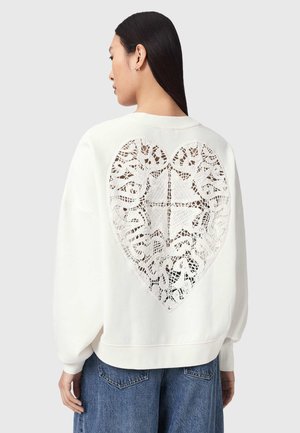Femme portant un sweat blanc avec une grande découpe en dentelle en forme de cœur dans le dos, associée à un jean bleu, tournée vers la gauche.