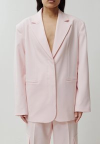 Femme portant un blazer oversize rose pâle et un pantalon assorti, debout devant un fond uni et neutre.