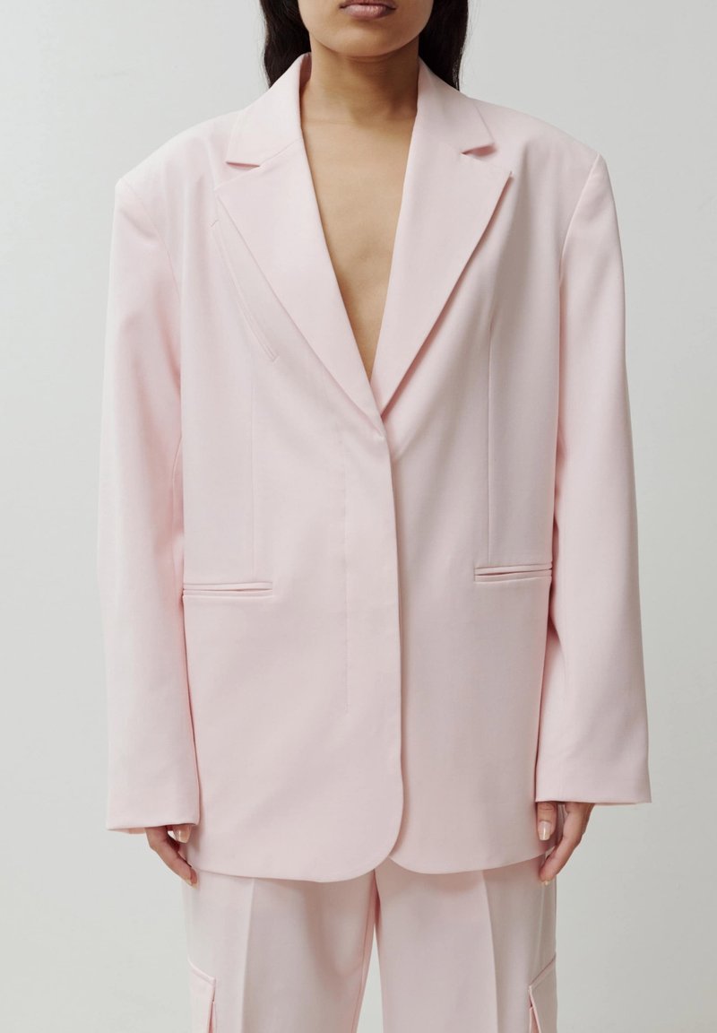 Femme portant un blazer oversize rose pâle et un pantalon assorti, debout devant un fond uni et neutre.
