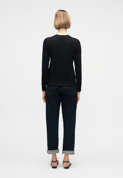 Γυναίκα με κοντά ξανθά μαλλιά που φοράει μαύρο μακρυμάνικο τοπ, σκούρο τζιν με γυρισμένα μανίκια και μαύρα σανδάλια, στέκεται με την πλάτη προς την κάμερα.