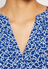 Tissu à motif bleu et blanc avec un décolleté en V. Le motif présente des formes irrégulières sur un fond bleu vif.