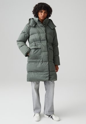 Groene pufferjas met een afneembare capuchon, een zandlopervormige taille, voorzakken en een gewatteerde textuur. Gekozen met lichtgrijze jeans en witte sneakers.
