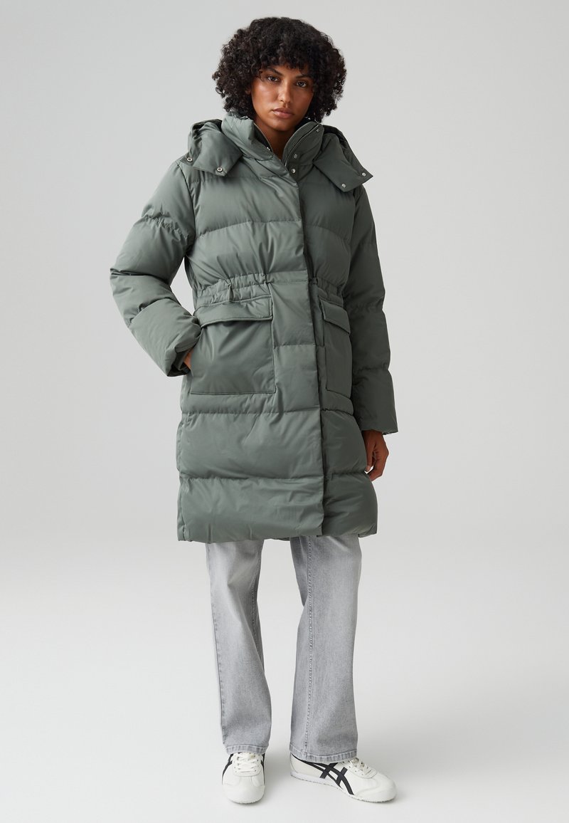 Cappotto puffer verde con cappuccio removibile, vita stretta, tasche anteriori e texture trapuntata. Indossato con jeans grigi chiari e sneaker bianche.