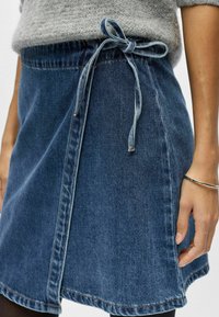 Jupe trapèze en denim avec panneau avant et nœud noué à la taille, portée avec un pull gris texturé et un bracelet argenté au poignet.