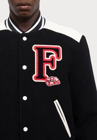 Zwarte wollen varsityjack met witte schouderaccenten, een prominente rode "F" geborduurd op de borst en lippenpatchdetail. Witte knopen en zakken.