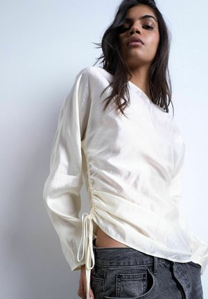 Femme portant un chemisier crème brillant à manches longues avec des liens sur les côtés, associé à un jean taille haute gris foncé sur un fond uni.