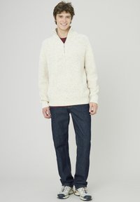 Un pull en tricot épais couleur crème avec un col zippé quart, associé à un jean bleu foncé et des baskets grises avec des accents colorés.