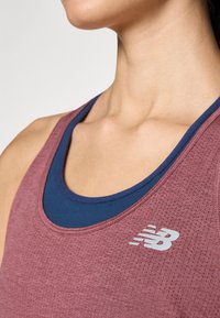 Maroon idrottstopp i ett strukturerat tyg, lager på lager över en marinblå topp. Har en reflekterande New Balance-logga på bröstet.