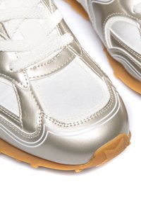 Srebrne i białe buty sportowe charakteryzują się gładką syntetyczną cholewką, teksturowanymi akcentami oraz podeszwą z gumy o wzorowanym gripie.