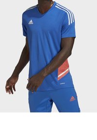 Modrý sportovní tričko s výstřihem do V, bílé třípruhové design na ramenou, červený akcent, a strukturovaná látka. Obsahuje logo Adidas.