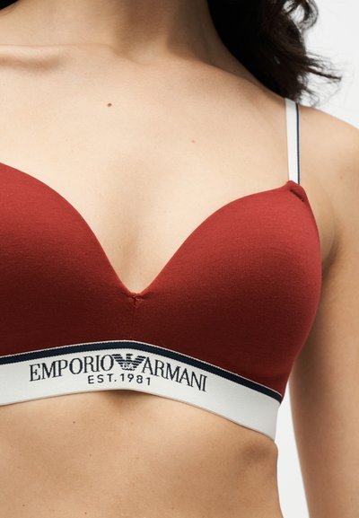 Γυναίκα που φοράει κόκκινο σουτιέν Emporio Armani με λευκή ελαστική λωρίδα και μαύρο λογότυπο, εμφανίζοντας τον άνω κορμό και τους ιμάντες σε λευκό φόντο.