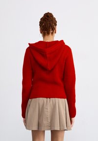 Rode gebreide hoodie met een textuurafwerking, voorzien van een kap en geribbelde zoom. Gecombineerd met een beige plooienrok.