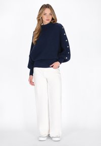 Maglione blu navy a maglia con texture a costine e bottoni lungo le maniche, abbinato a pantaloni bianchi a gamba larga e sneakers bianche.