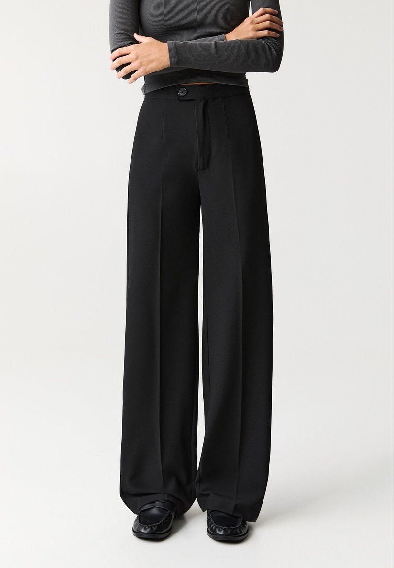 Femme portant un pantalon noir taille haute à jambes larges, des chaussures noires, et un haut moulant à manches longues gris foncé, bras croisés.