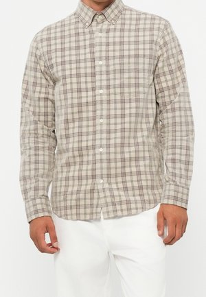 Mann trägt ein beige-grau kariertes Hemd mit langen Ärmeln und Kragen, kombiniert mit weißer Hose, steht vor einfachem Hintergrund.