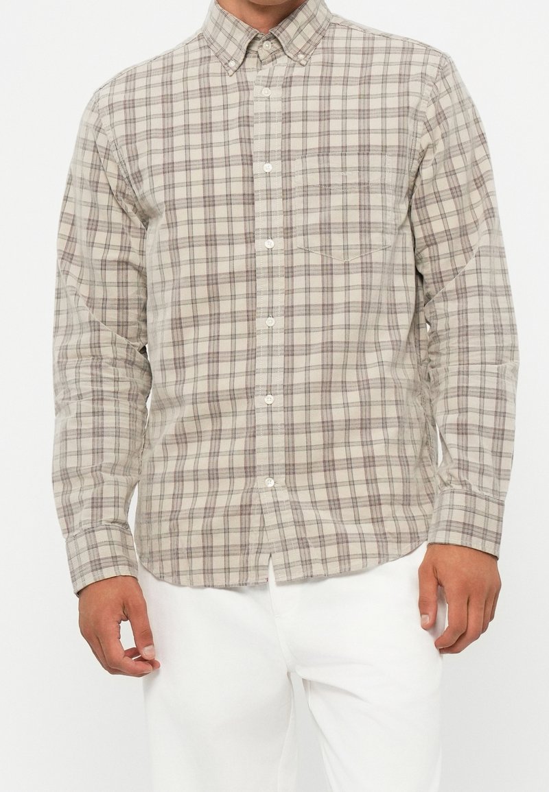 Homme portant une chemise à manches longues à carreaux beige et gris avec col, assortie à un pantalon blanc, debout devant un fond uni.