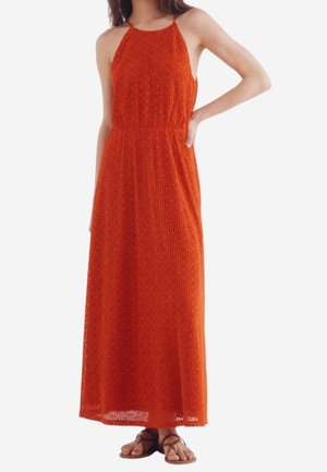 Femme portant une robe orange en crochet sans manches, longue jusqu'aux chevilles, avec un col halter et des sandales plates marron.