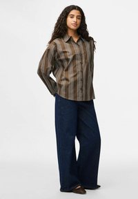 Donna con lunghi capelli ricci, che indossa una camicia a righe marrone con bottoni, jeans a gamba larga blu scuro e scarpe a punta marroni, in piedi davanti a uno sfondo bianco.