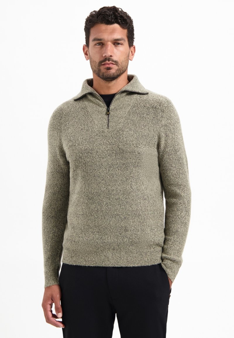 Man in een gemeleerd lichtgrijs kwart-rits sweater en zwarte broek, staand met één hand in zijn zak tegen een witte achtergrond.