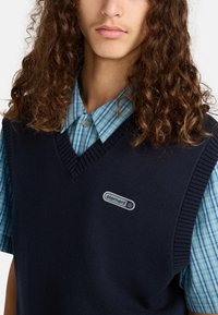 Marine gebreide vest met geribde randen, gedragen over een lichtblauw en wit geblokt shirt. Beschikt over een logo-opdruk op de borst.