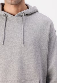 Haut à capuche gris en coton doux. Il dispose d'une poche frontale et de cordons de serrage ajustables. La texture est lisse et légèrement polaire.