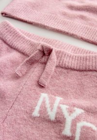 Hellrosa gestricktes Set mit einer Mütze und passendem Unterteil. Verfügt über einen Kordelzug und weiße "NYC"-Buchstaben auf dem Stoff. Weicher Stoff.
