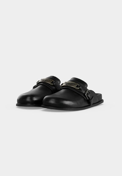 Μαύρα δερμάτινα clog slip-on με κλειστά δάχτυλα, διακοσμητική μεταλλική αγκράφα και επίπεδη σόλα σε απλό ανοιχτό φόντο.