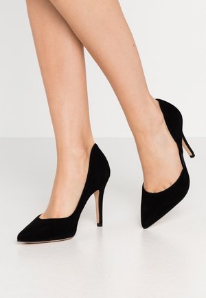 Jambes portant des escarpins en daim noir à bout pointu avec des talons aiguilles fins, posées sur un sol lisse et clair.
