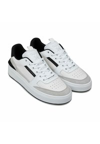 Witte en grijze sneakers van leer en suède, met geperforeerd teenstuk, zwarte accenten en witte veters op een brede rubberen zool.