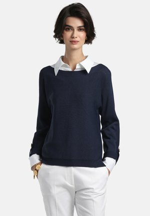 Tumši zils adīts džemperis ar plašu kakla izgriezumu un gariem piedurknēm, kārtots virs baltas kreklblausas ar apkakli. Piedurkņu malās ir zelta pogu akcenti.