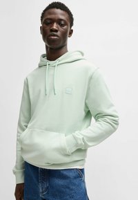 Light green hoodie with a front pocket, adjustable drawstrings, and logo patch. Paired with blue denim shorts, smooth fabric texture.