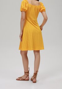 Robe jaune aux manches bouffantes, avec des détails en dentelle et un décolleté carré, associée à des sandales marron à lacets.