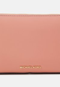 Vaalea vaaleanpunainen nahkapussi, jossa on kultainen vetoketju ja "Michael Kors" -brändinimi kultakirjaimin keskitettynä etupuolelle.