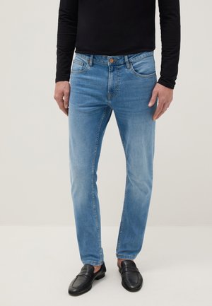 Hombre vistiendo jeans ajustados de color azul claro, camisa negra de manga larga y mocasines de cuero negro, de pie contra un fondo liso.