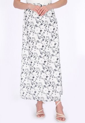 Kobieta ubrana w białą, sięgającą kostek spódnicę z czarnym kwiatowym wzorem i beżowe, wysokie sandały na paski, z rękami złożonymi na wysokości talii.