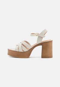 Sandali platform color beige chiaro con tacco in legno, caratterizzati da più strap incrociati, una cinghia alla caviglia con una fibbia dorata e una texture liscia.