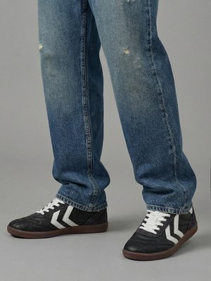 Person trägt abgenutzte blaue Jeans und schwarze Sneaker mit weißen Chevron-Streifen und steht auf einem grauen Boden.