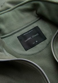 Veste texturée verte avec une fermeture éclair argentée et une étiquette noire à l'intérieur du col indiquant « Qualité Premium Ombre Exmio Taille M ».