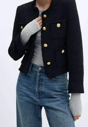 Osoba ubrana w czarną, teksturowaną kurtkę z złotymi guzikami na szarej, długorękawowej koszuli i niebieskich, wysokich dżinsach, z ręką częściowo zakrytą przez rękaw.