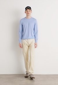 Suéter de manga longa azul claro, calças de cor creme, sapatilhas cinzentas e um boné preto com texto branco, usados por uma figura masculina em pé.