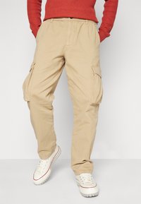 Pantalones de cargo color beige con bolsillos laterales, hechos de una resistente tela de algodón. Tienen un corte relajado y piernas rectas, combinados con zapatillas blancas.