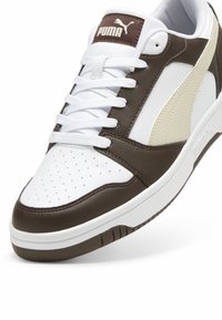 Białe i brązowe buty sportowe wykonane ze skóry i materiałów syntetycznych, z perforowanym czubkiem, białymi sznurowadłami i gumową podeszwą. Logo PUMA na języku.