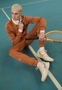 Oranžni trenirka z rdečimi črtami, beli ovratnik; sedi na modrem igrišču. Drži teniško loparico, nosi pastelne Nike superge.