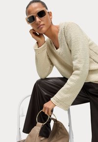 Maglione lavorato a maglia beige con scollo a V e motivo testurizzato, abbinato a pantaloni di pelle marrone scuro. L'accessorio include una borsa beige con manici circolari dorati.