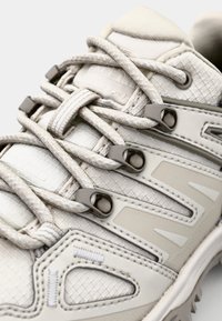Light-colored athletic shoe featuring textured synthetic material, prominent lacing system, and lightweight midsole with unique cut-out design details.