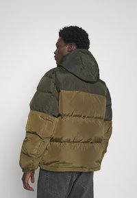 Chaqueta acolchada con capucha, que presenta paneles en color oliva y verde oscuro, textura acolchada, puños elásticos y un corte relajado.