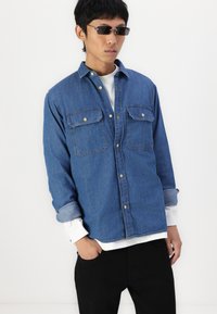 A denim shirt in medium blue with two chest pockets, buttoned front, and rolled-up sleeves over a white top. Black pants complete the look.