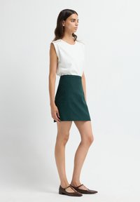 Top bianco senza maniche con decorazioni eleganti sulle spalle, abbinato a una minigonna fitted verde scuro e scarpe flat a T color marrone.