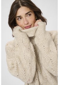 Svijetlosmeđi pulover od pletene vune s visokim rebrastim turtleneck-om i slobodnim krojem. Izrađen od mekog, strukturiranog materijala s otvorenim uzorkom.