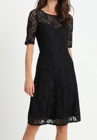 Robe en dentelle noire avec des manches courtes, un corsage ajusté et une jupe évasée. Le motif complexe de la dentelle comprend des détails floraux.